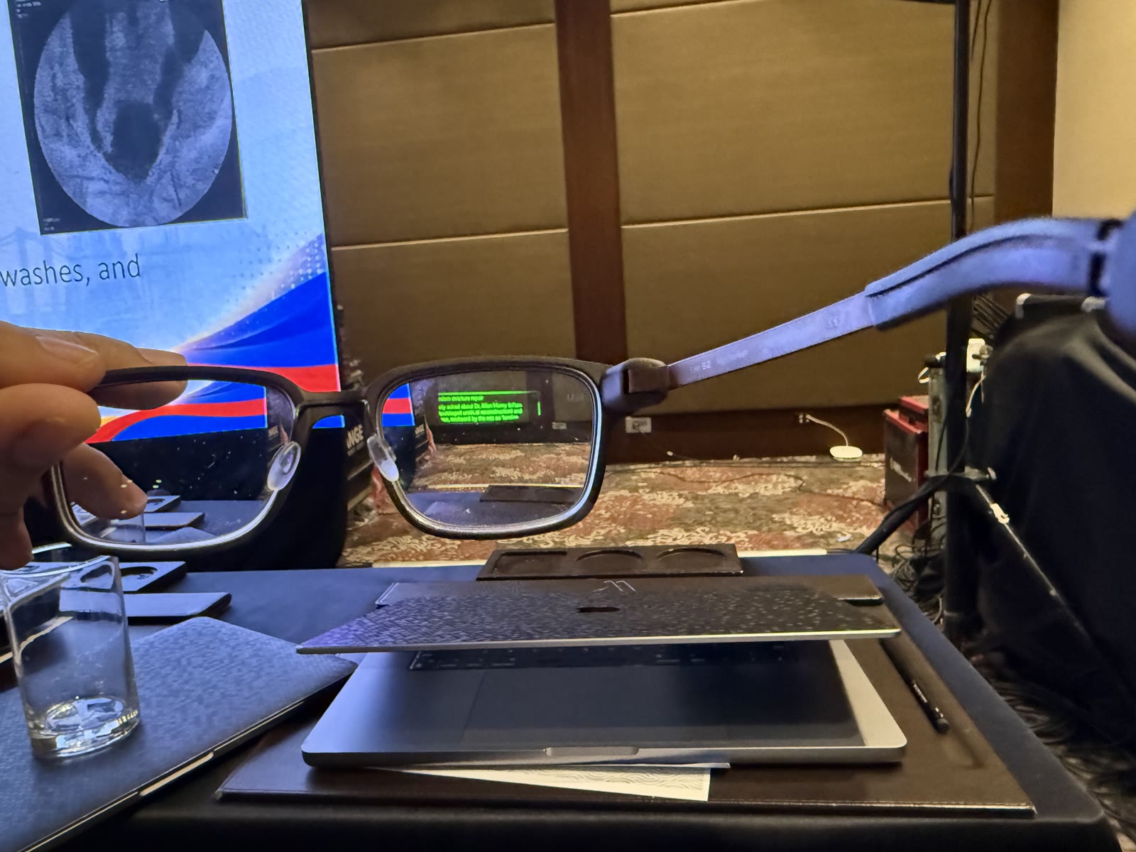 A hand holds the Even G2 glasses in front of a lecture screen, with several lines of bright green text visible through one lens.