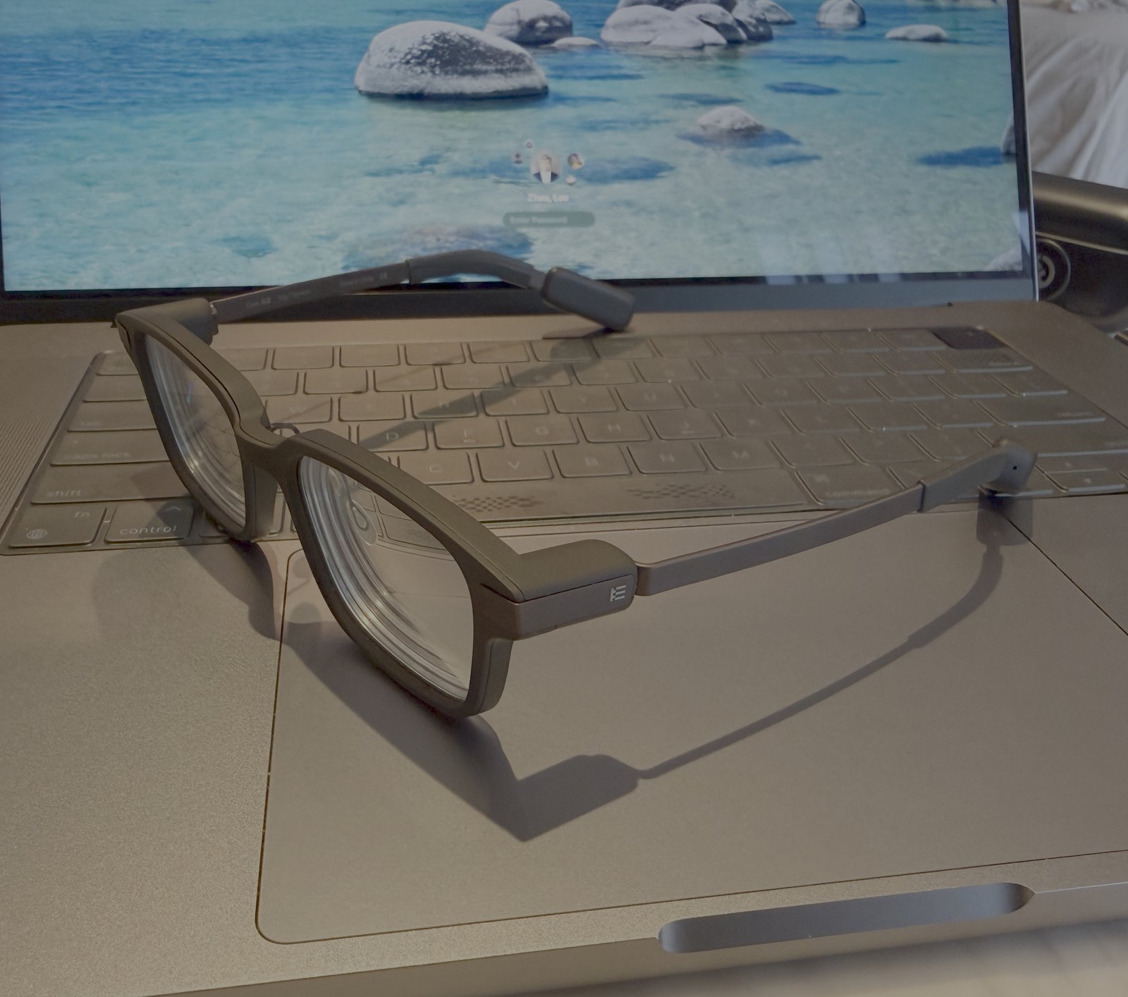 A pair of Even G2 glasses, dark matte frame with the temples open, resting on a laptop keyboard in front of a screen showing turquoise water and boulders.