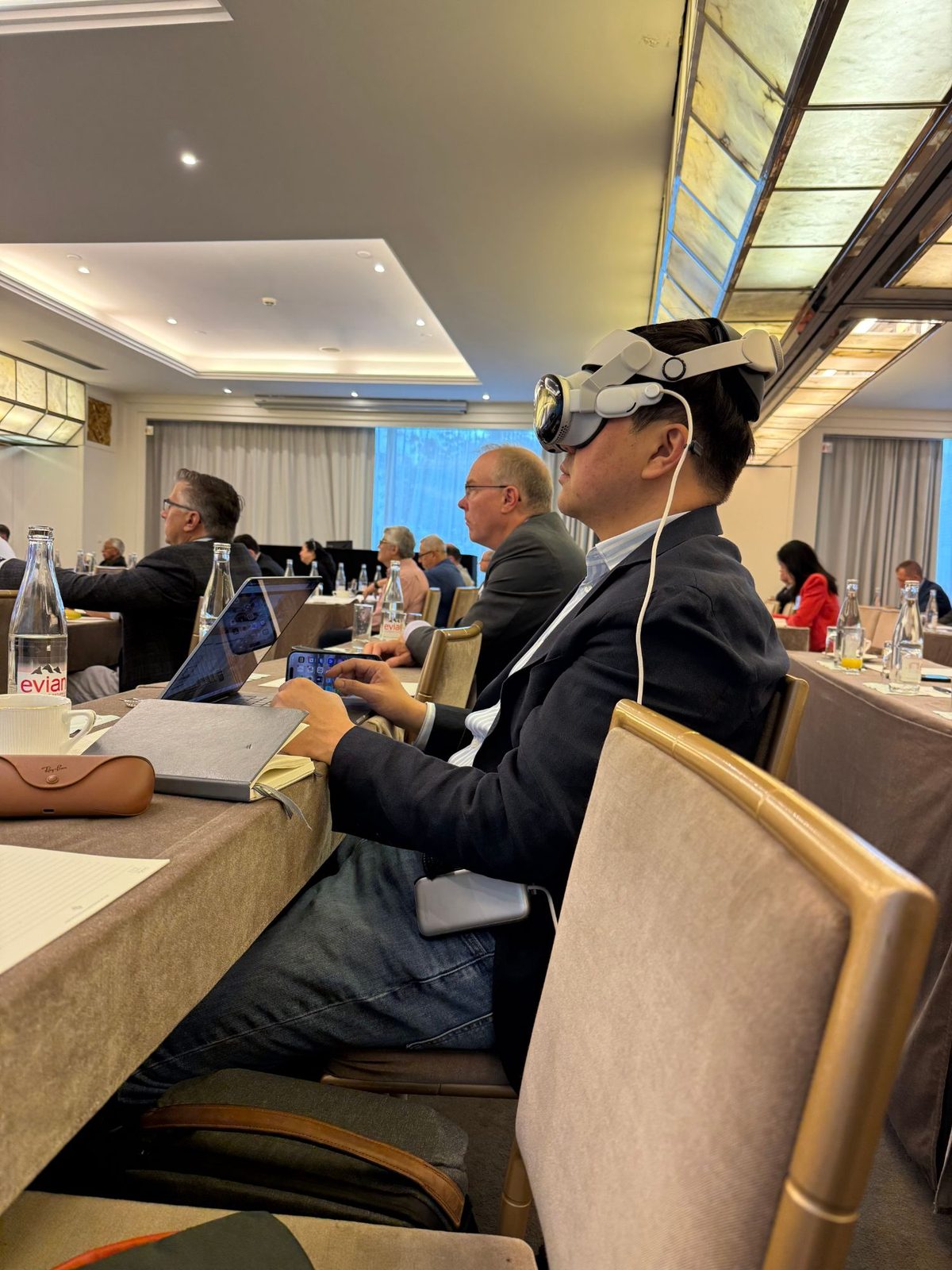 Lee Zhao seated at a conference table wearing an Apple Vision Pro headset, working at a laptop among other attendees in a meeting room